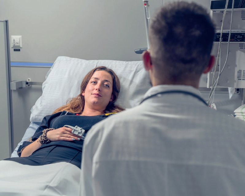 Patiente allongée dans un lit et un soignant de dos, face à la patiente