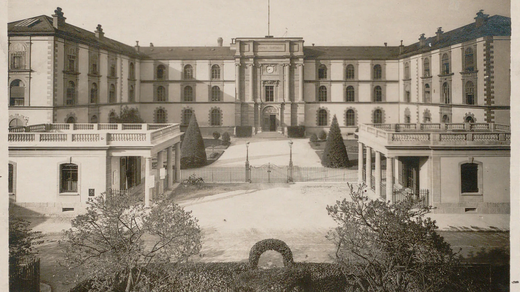 Entrée monumentale surplombée d'une horloge, deux pavillons au premier plan.