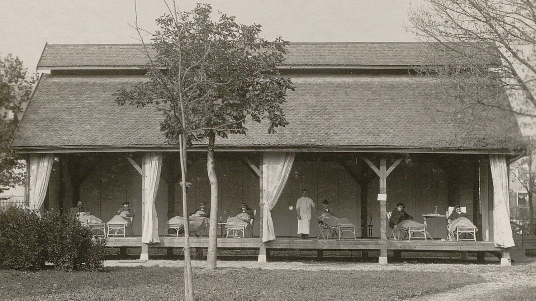 Pavillon sans murs, rideaux ouverts, un infirmier et plusieurs malades dans des chaises-longues.