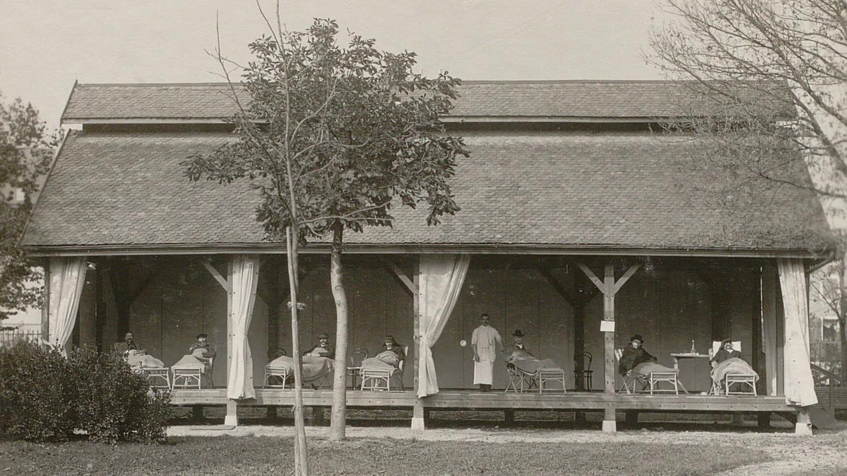 Pavillon sans murs, rideaux ouverts, un infirmier et plusieurs malades dans des chaises-longues.