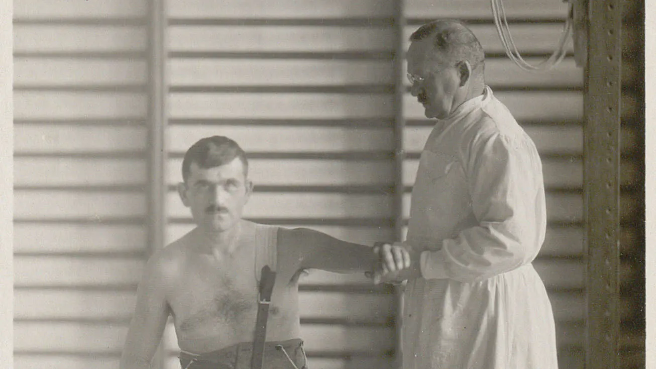 Homme à torse nu, assis sur une chaise, un physiothérapeute lui tient le bras.