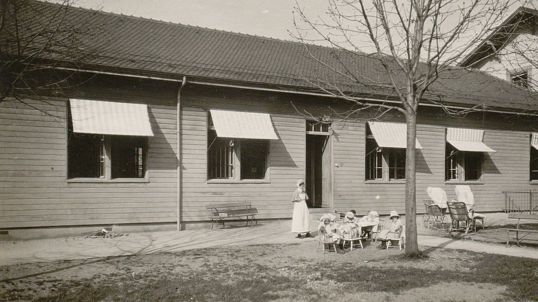 Dans le jardin devant le pavillon, des jeunes enfants sont assis sur des petites chaises, surveillés par une infirmière.
