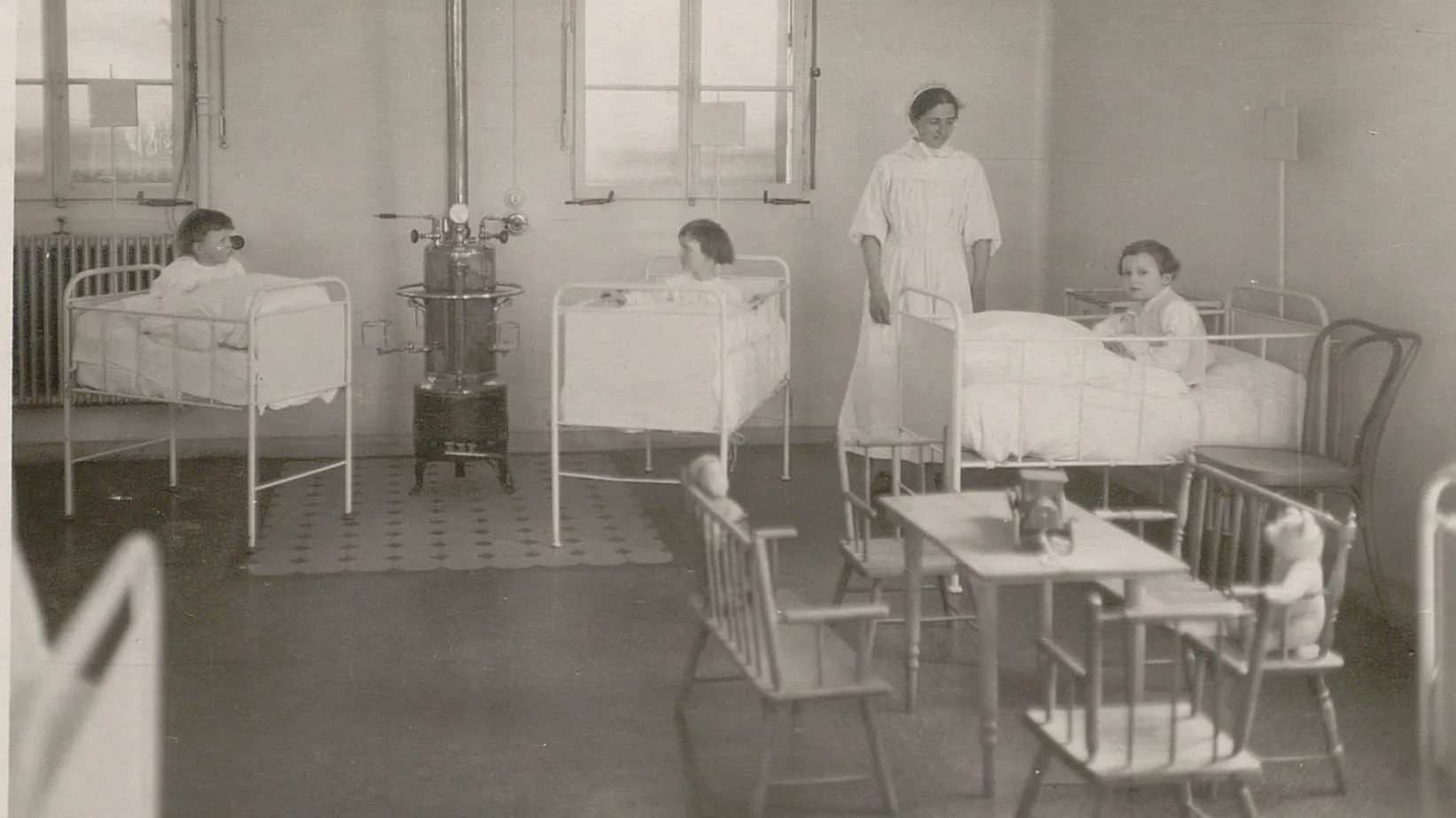 Trois jeunes enfants dans des lits à barreaux, surveillés par une infirmière. Un coin jeu avec un nounours.