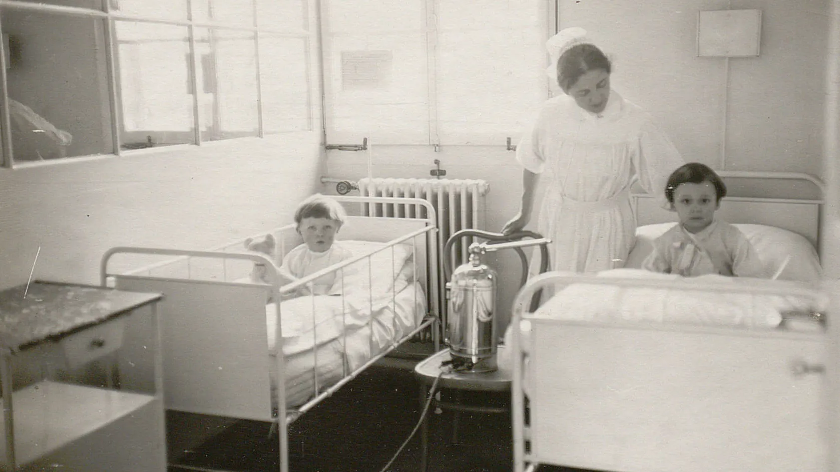 Deux jeunes enfants dans des lits, surveillés par une infirmière.