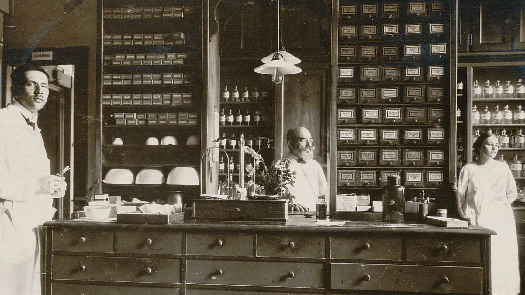 Trois personnes en blouse blanche devant des meubles remplis de pots et bouteilles, nombreux tiroirs.
