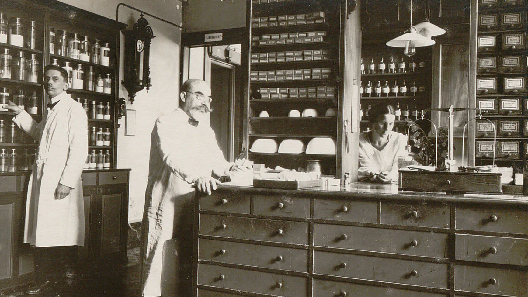Trois personnes en blouse blanche devant des meubles remplis de pots, bouteilles et tiroirs.