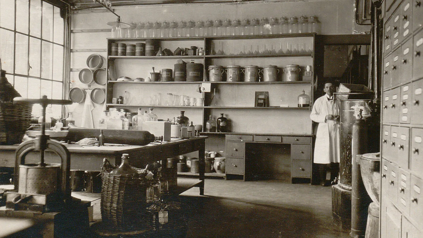 Salle équipée d'étagères remplies de bouteilles et contenants, d'une table de travail, d'une presse.