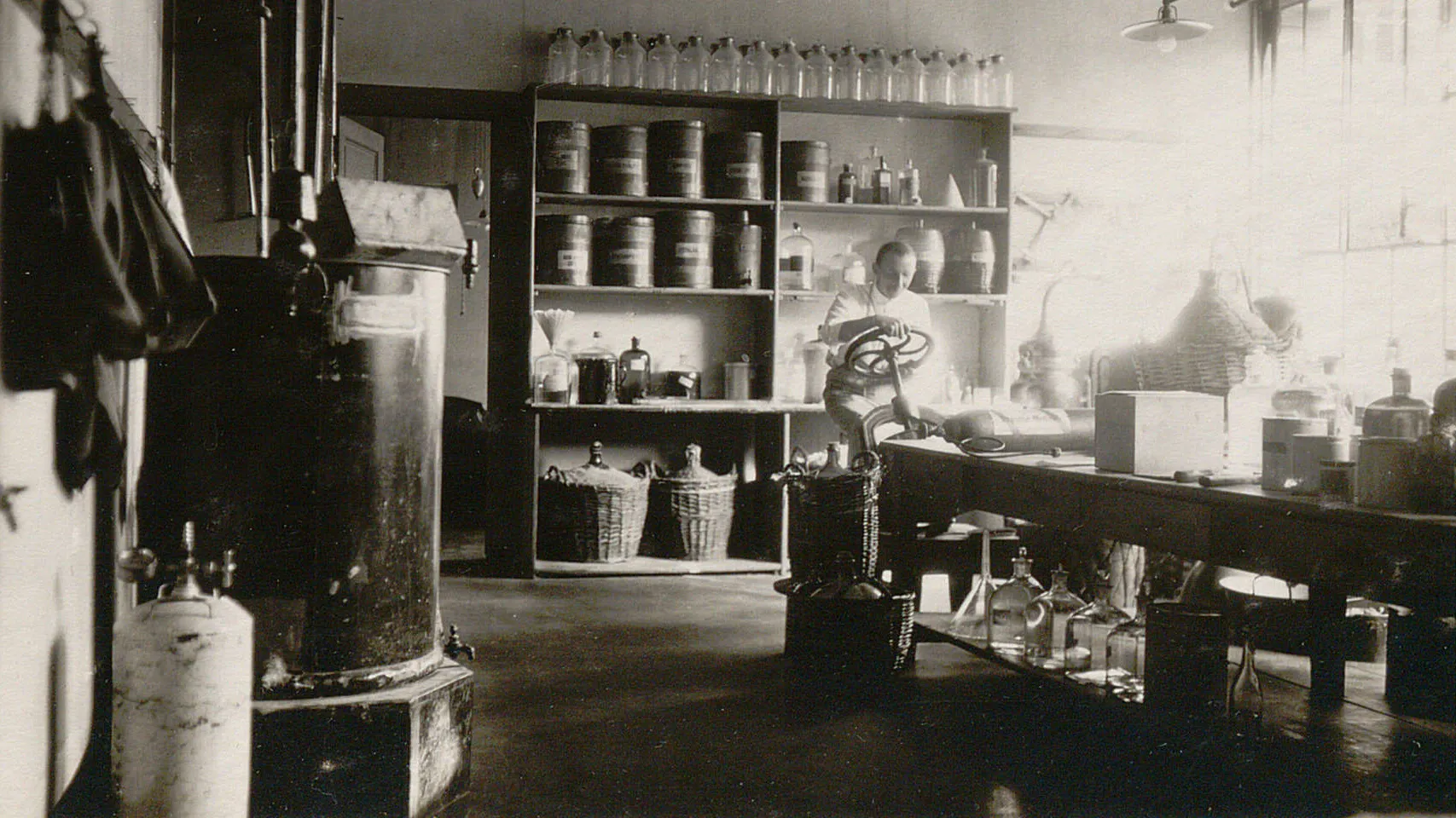Salle équipée d'étagères remplies de bouteilles et contenants, d'une table de travail, d'un pressoir.