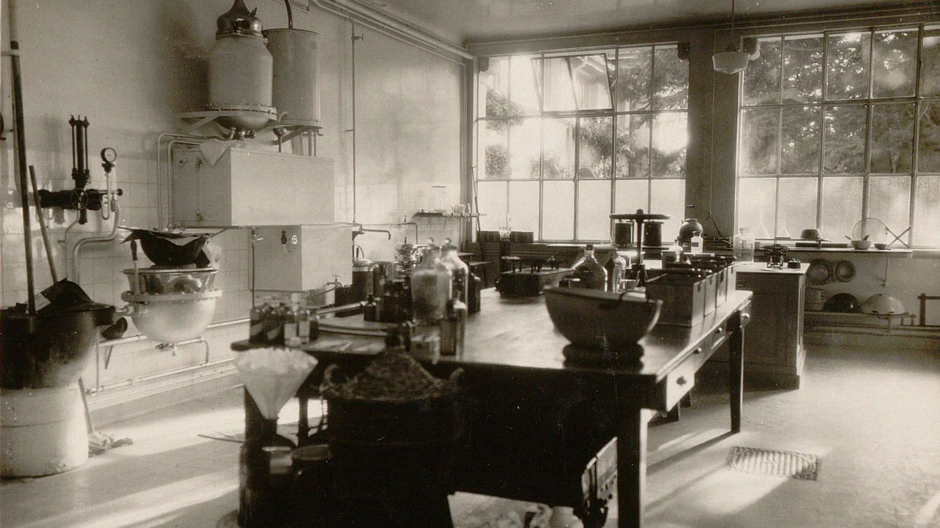 Salle avec des grandes fenêtres, une table au centre avec de nombreuses bouteilles, à gauche des bassines et une chaudière.