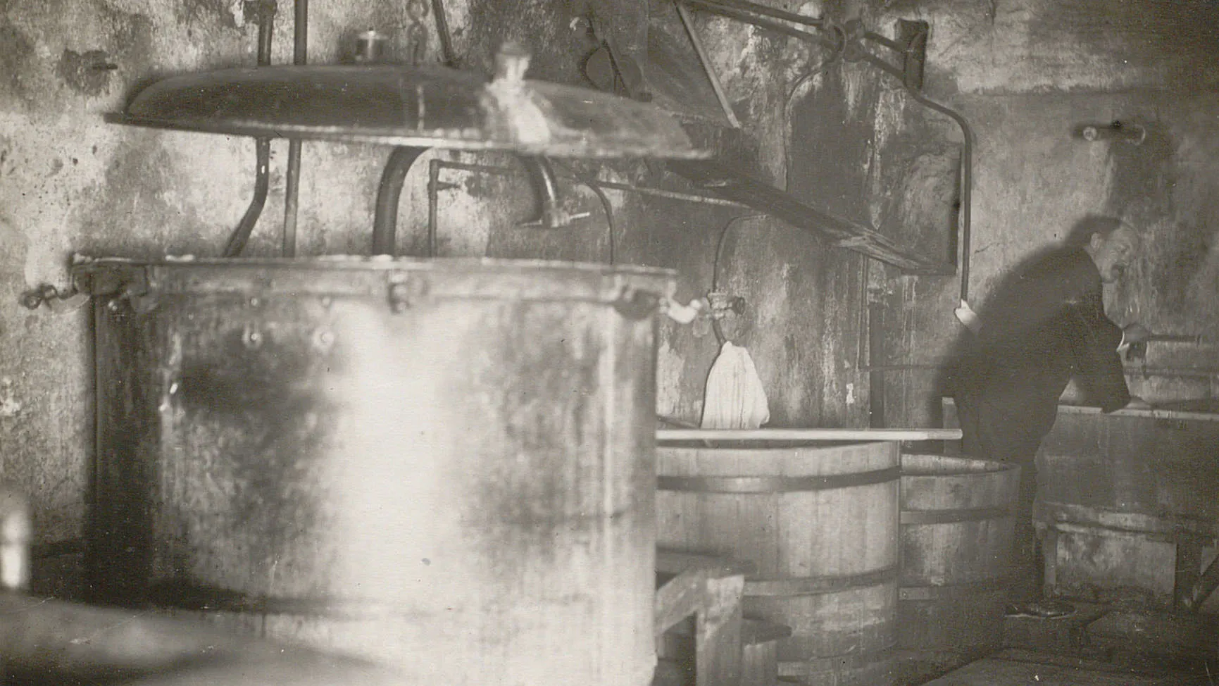 Plusieurs grandes cuves en métal ou en bois dans une pièce sombre, un homme se penche sur un évier au fonds de la pièce.