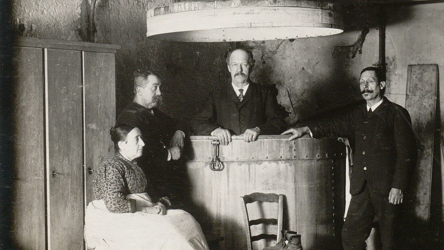 Un homme debout dans une grande cuve, deux autres hommes à gauche et à droite, une femme assise.