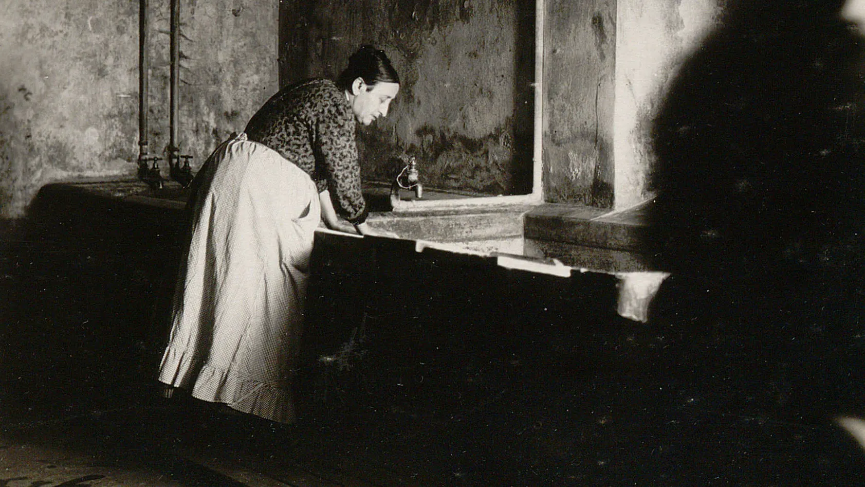 Une femme avec tablier se penche sur un lavoir muré.