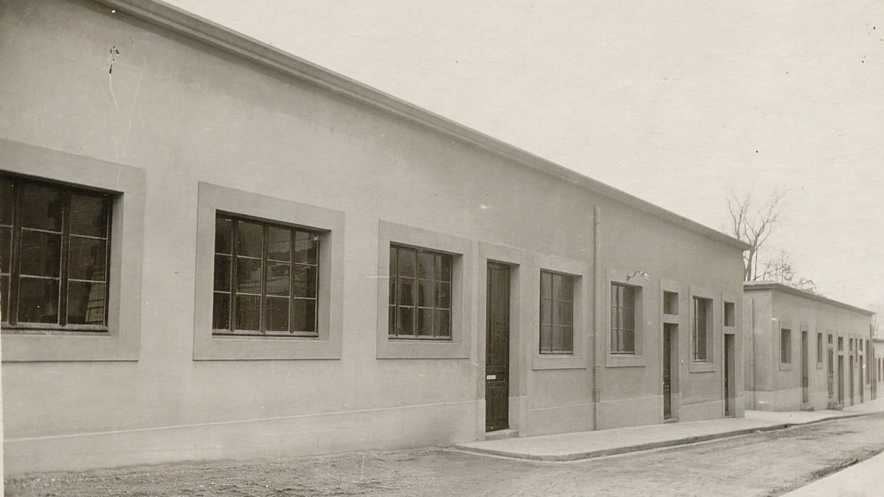Une rangée de bâtiments bas au toit plat, avec des nombreuses portes et fenêtres sans volets, devant un chemin en terre.