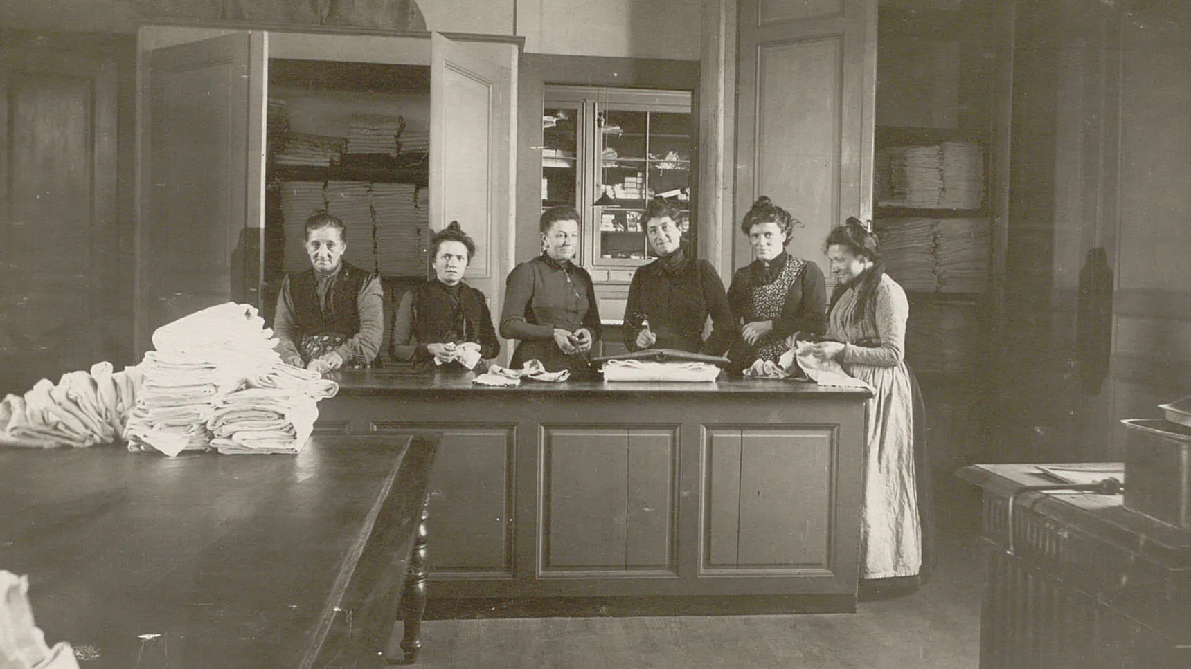 Six femmes derrière un comptoir en train de plier du linge qui sera rangé dans les armoires derrière elles.