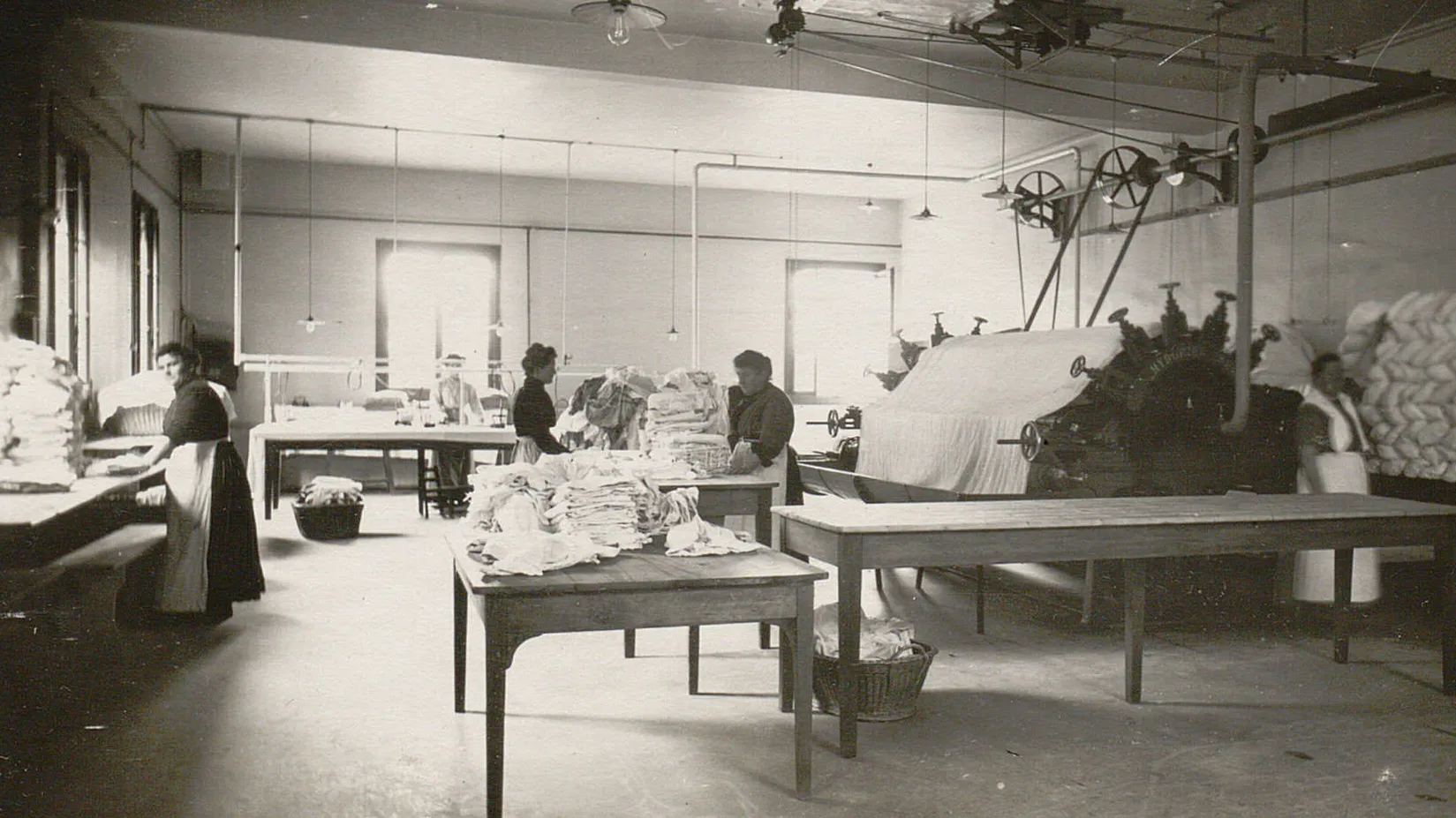 Une grande calandre, plusieurs tables avec des tas de linge plié, quelques femmes ouvrières.