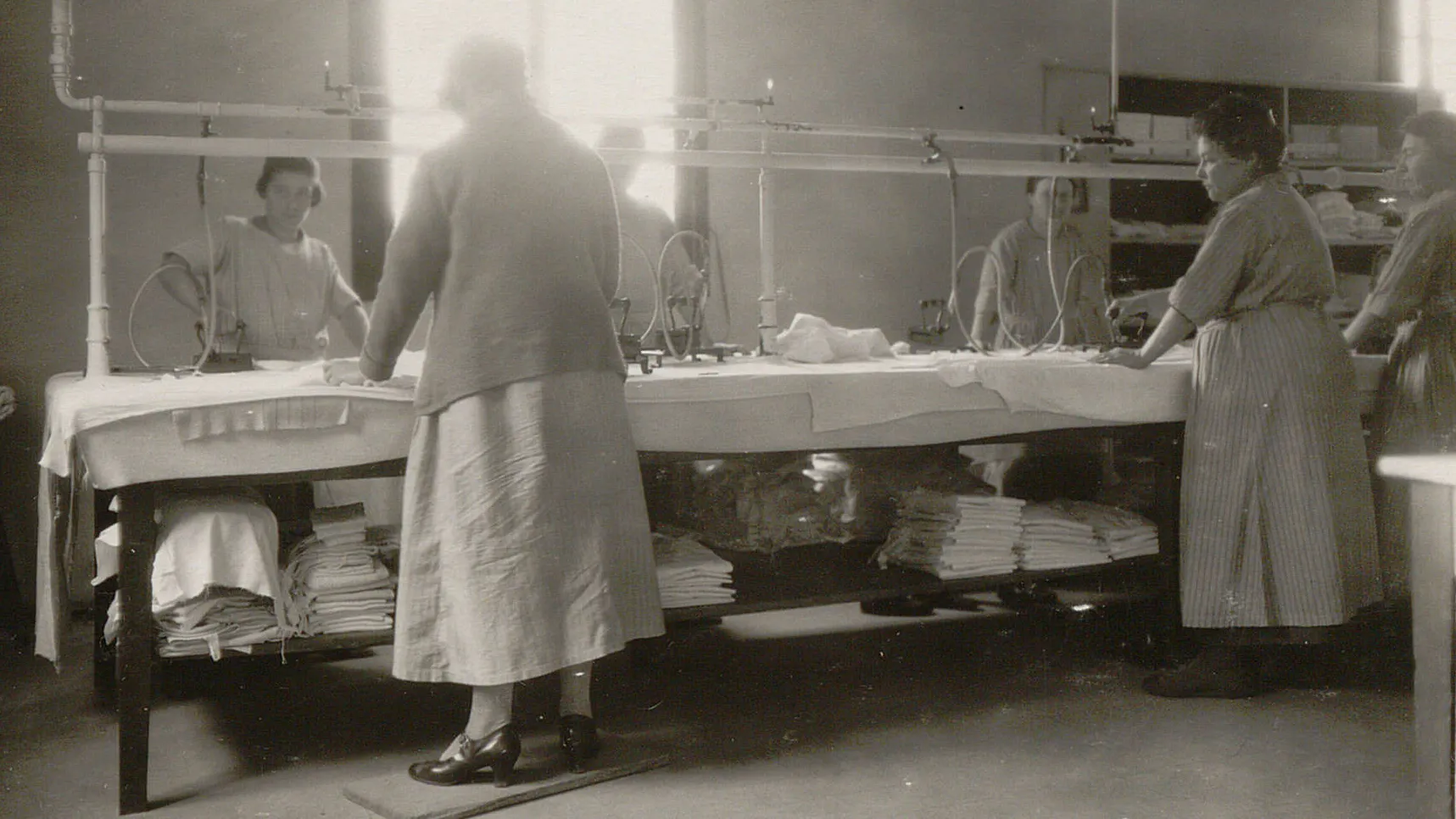 Six femmes autour d'une table couverte d'un molton, elles utilisent des fers à repasser à vapeur alimentés par des tuyaux.