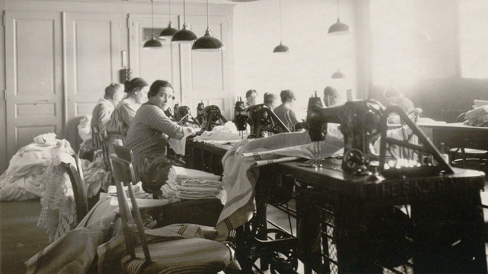 Rangée de tables avec des machines à coudre, plusieurs femmes y travaillent sur du linge à repriser.