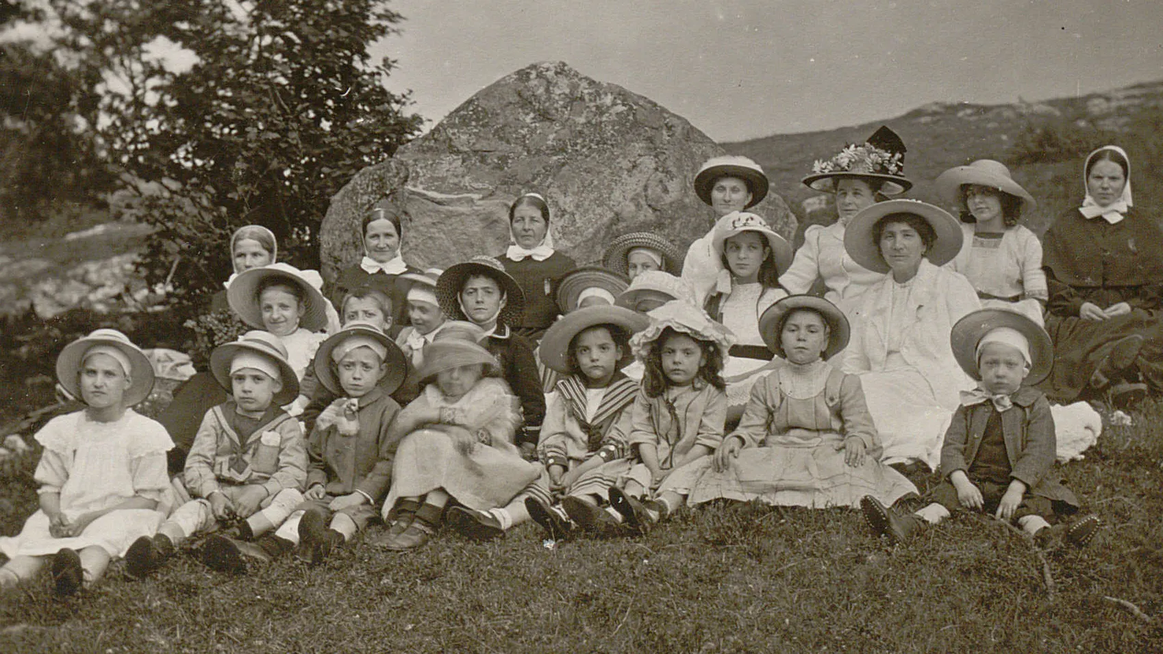 Groupe d'enfants en habits de dimanche assis devant un rocher, avec quelques diaconesses.