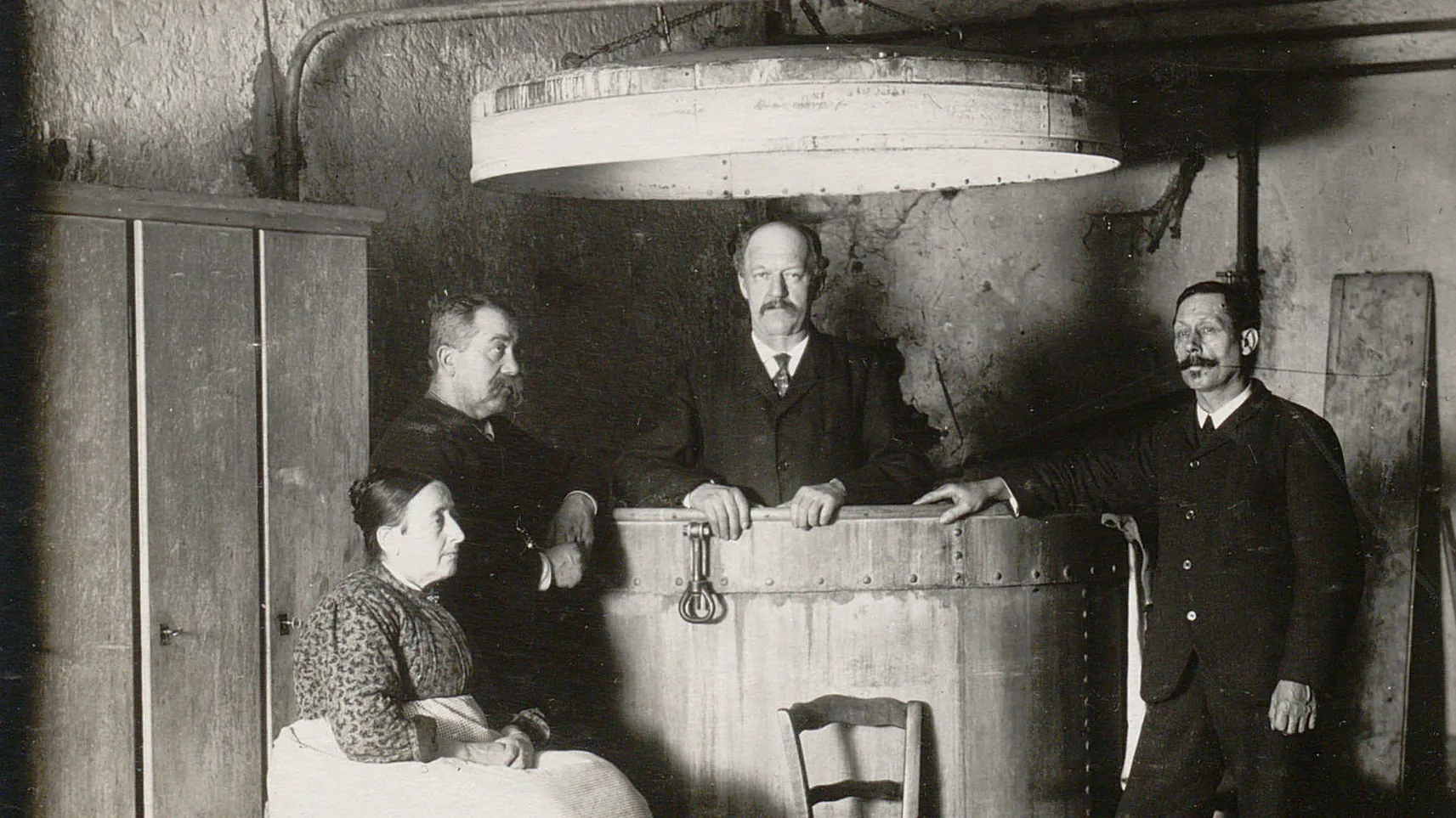 Un homme debout dans une grande cuve, deux autres hommes à gauche et à droite, une femme assise.