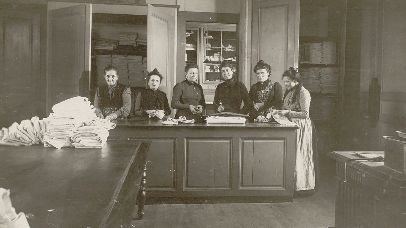 Six femmes derrière un comptoir en train de plier du linge qui sera rangé dans les armoires derrière elles.