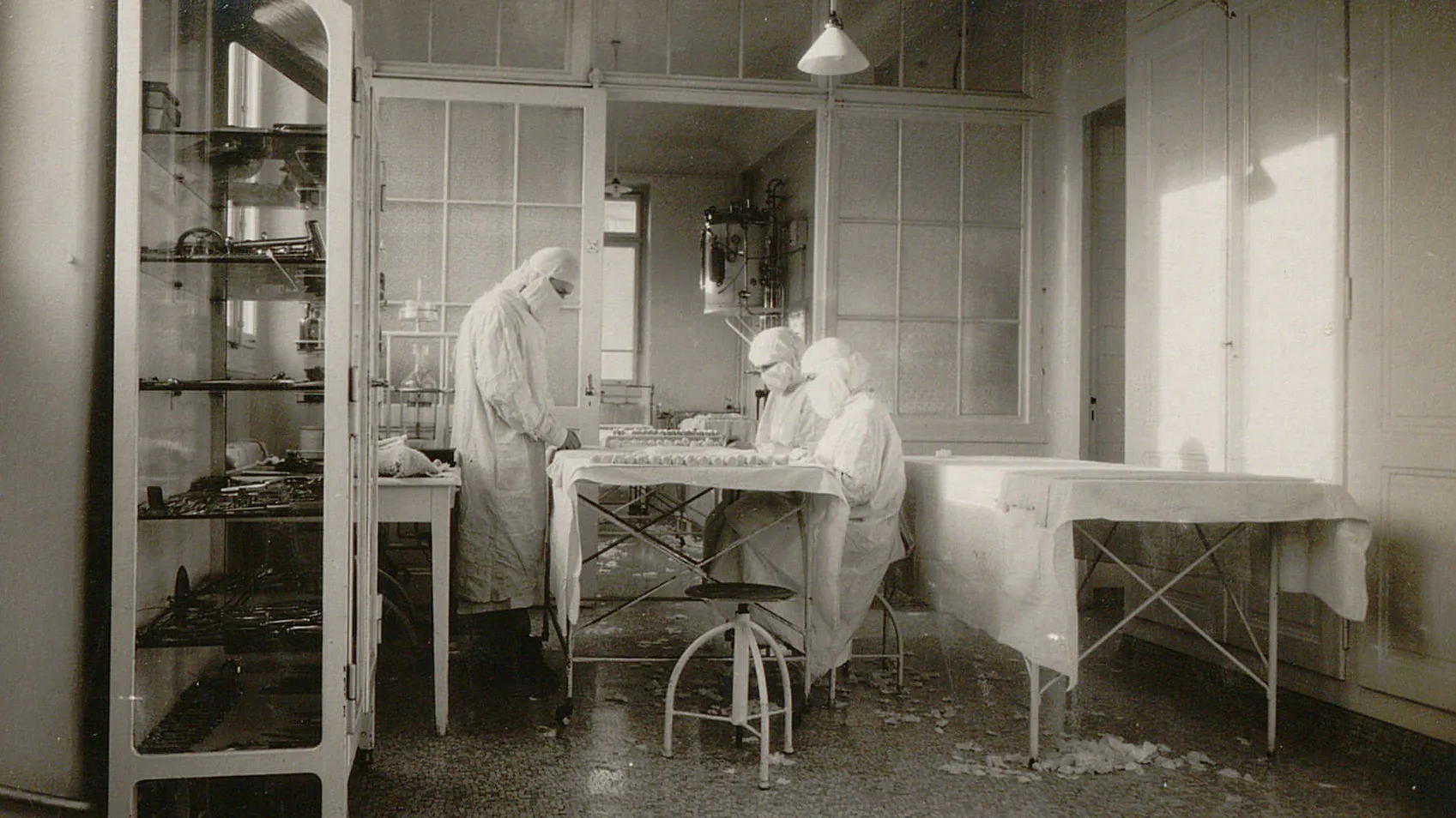Trois personnes avec masque et charlotte préparent des rouleaux de pansements ou de bandages.