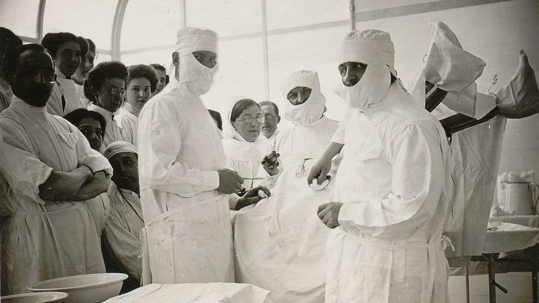 Trois chirurgiens avec charlotte et couvre-barbe ainsi qu'un public nombreux entourent un patient.