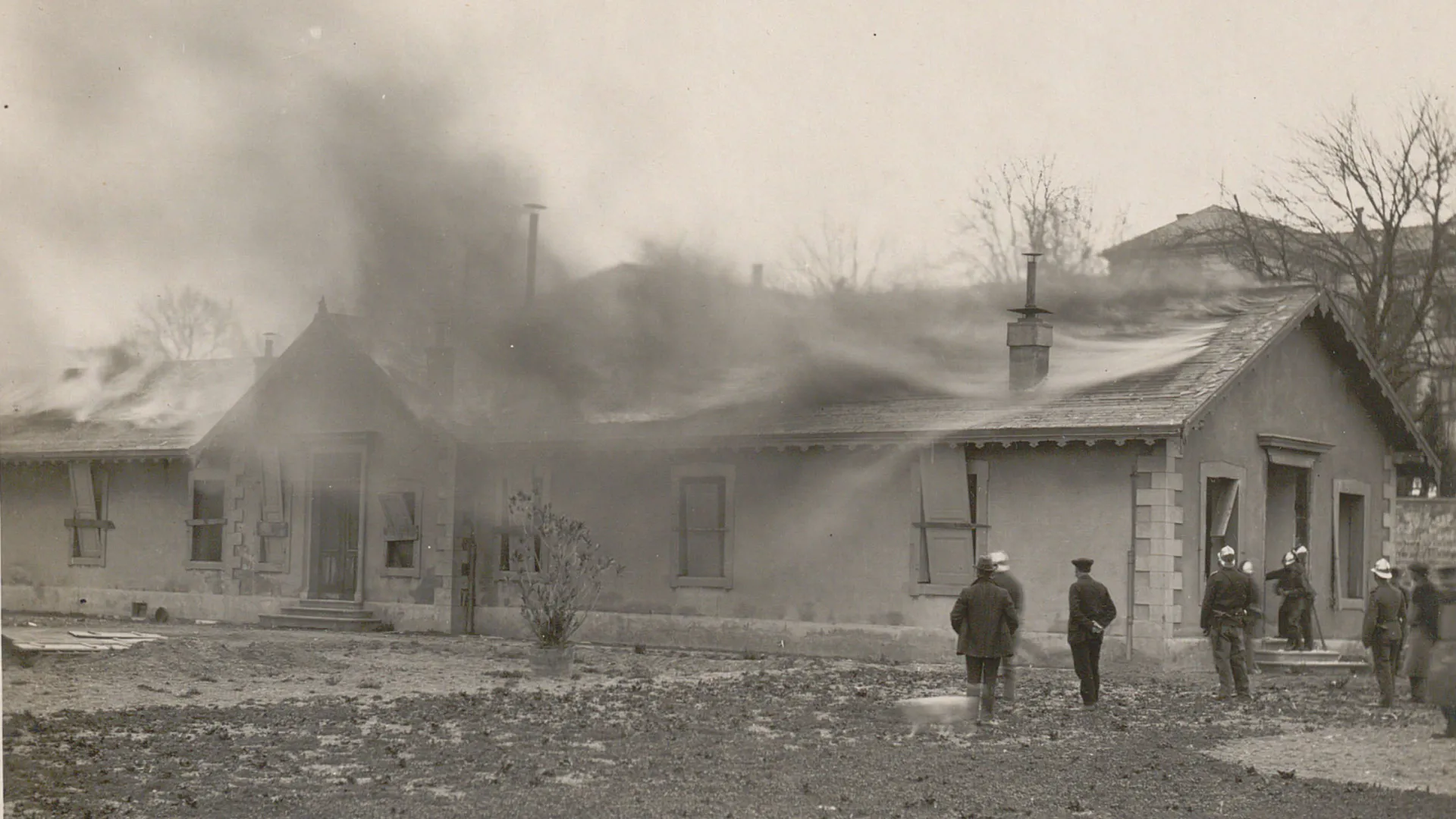 Le toit du pavillon est en feu, beaucoup de fumée. Des pompiers observent la scène.