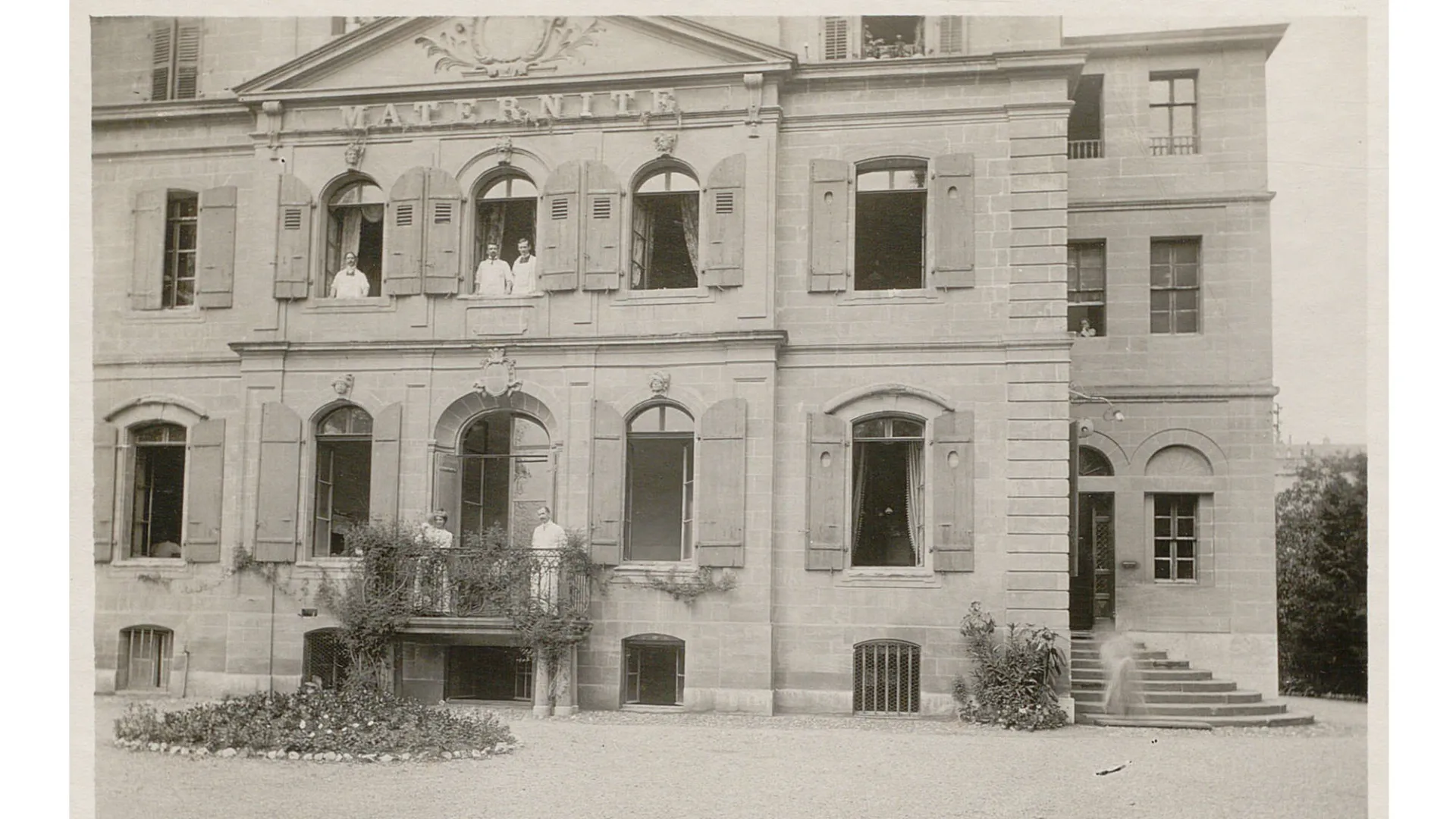 Bâtiment à plusieurs étages avec une inscription "Maternité". Des personnes en blouse blanche sont aux fenêtres.