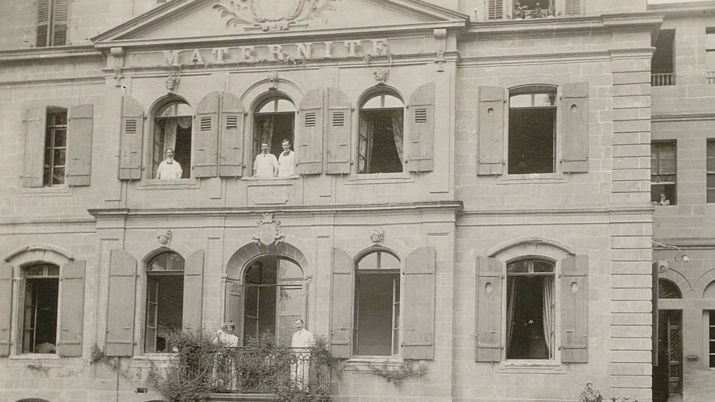 Des personnes en blouse blanche regardent par les fenêtres d'un bâtiment marqué "Maternité".