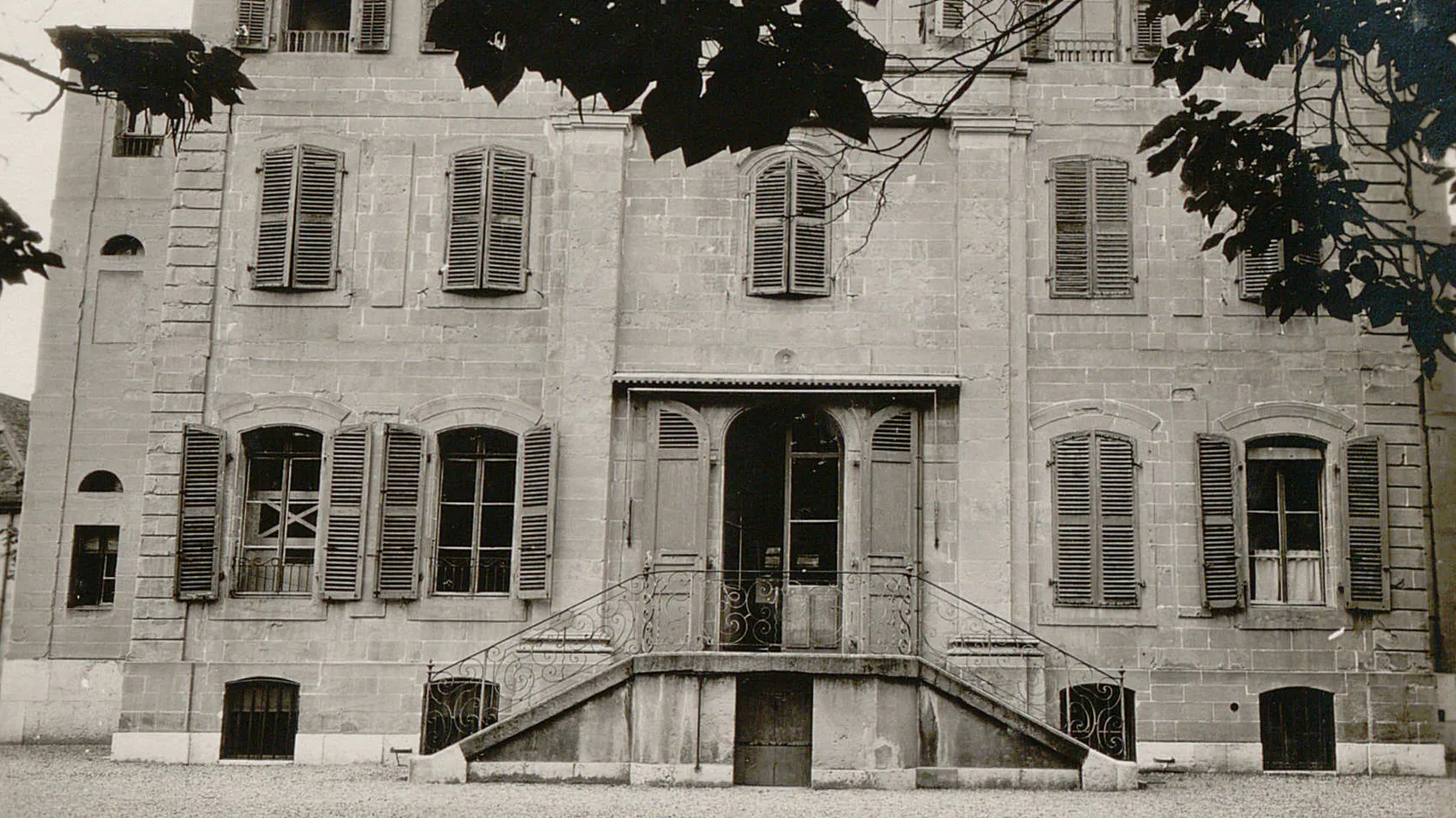 Bâtiment à plusieurs étages, des grandes fenêtres à volets. Un double escalier mène vers une grande porte au centre.