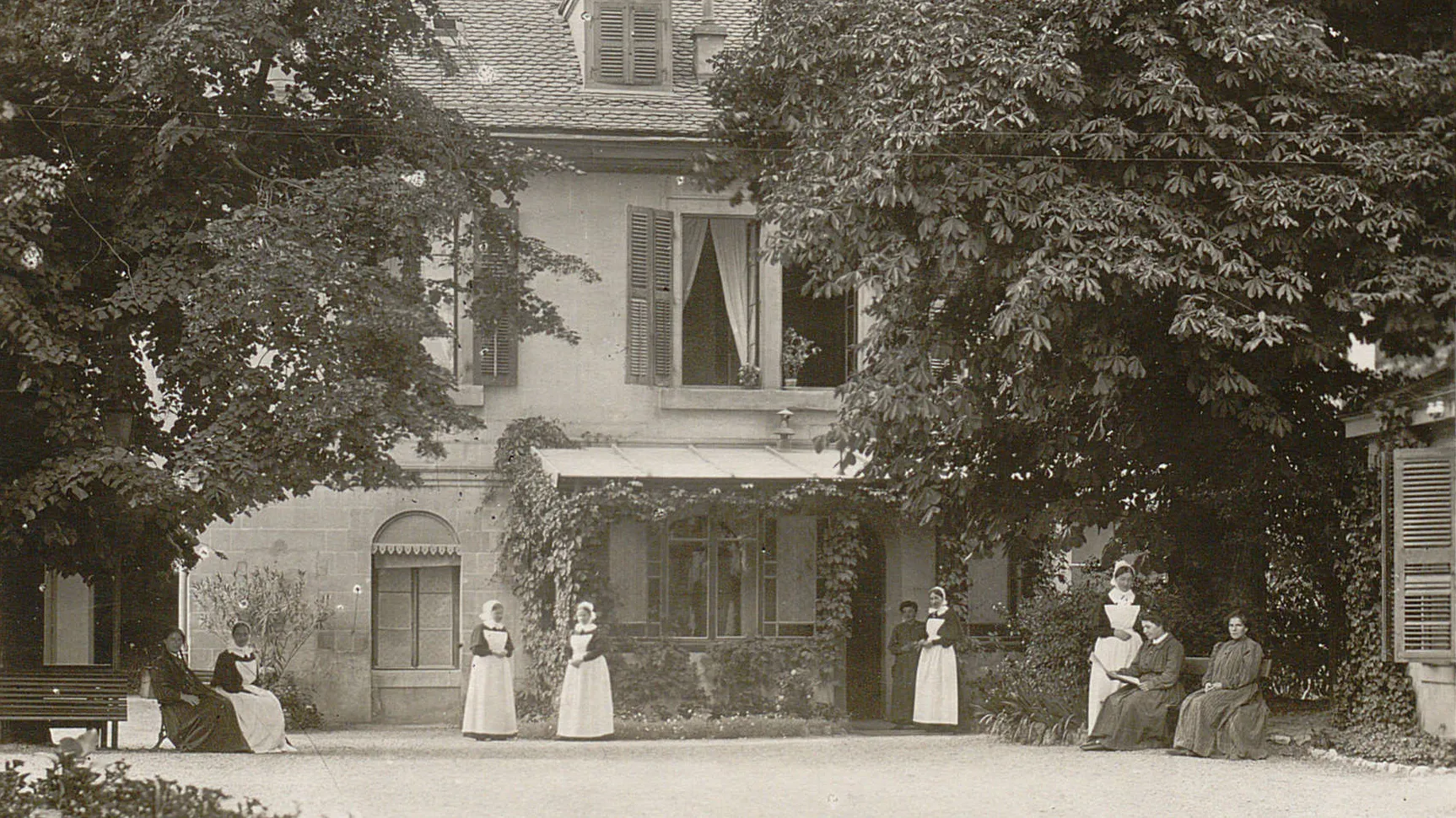 Une cour avec des grands arbres, plusieurs femmes sont assises sur des bancs. Des diaconesses sont près d'elles.