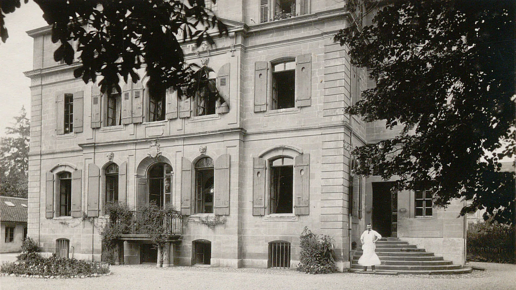 Bâtiment à plusieurs étages. Un escalier latéral mène vers une porte sur la droite. Un homme en blouse blanche s'y trouve.