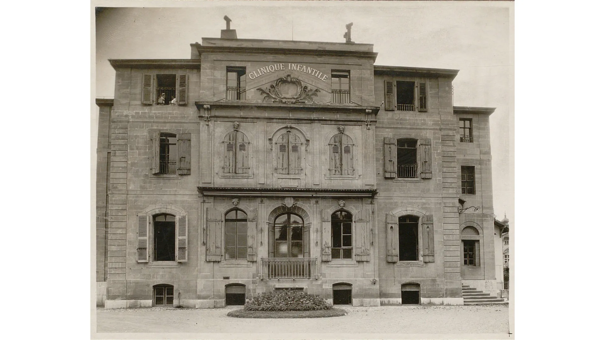 Bâtiment à plusieurs étages avec une inscription "Clinique infantile". Un balcon au centre, un escalier à droite.