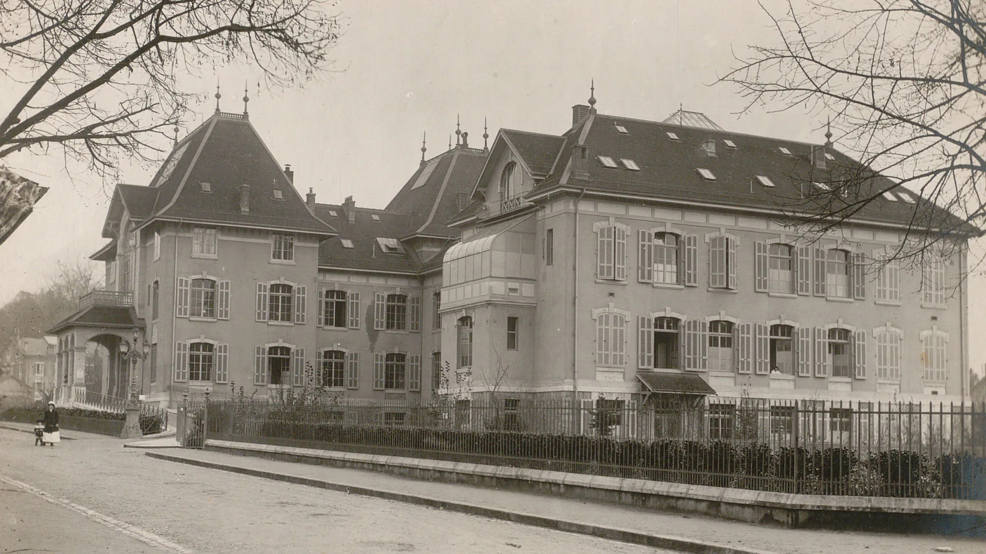 Vue sur la Maternité, un bâtiment de trois étages, depuis la route où une femme et un enfant se promènent.