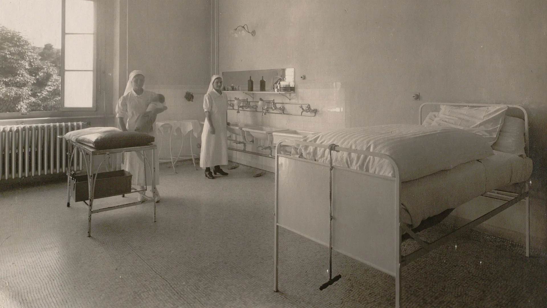 Une infirmière avec un bébé dans les bras dans une salle avec un lit d'hôpital très haut, des lavabos, un chandelier.