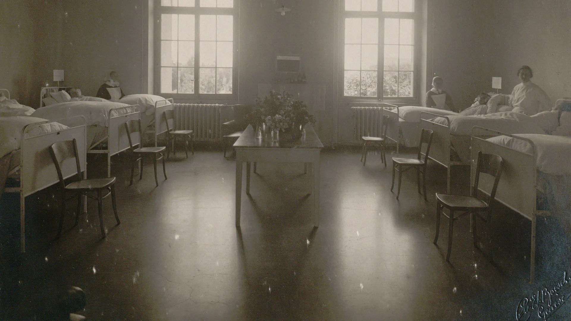 Chambre à six lits, plusieurs patientes et diaconesses, une table au centre avec un bouquet de fleurs.