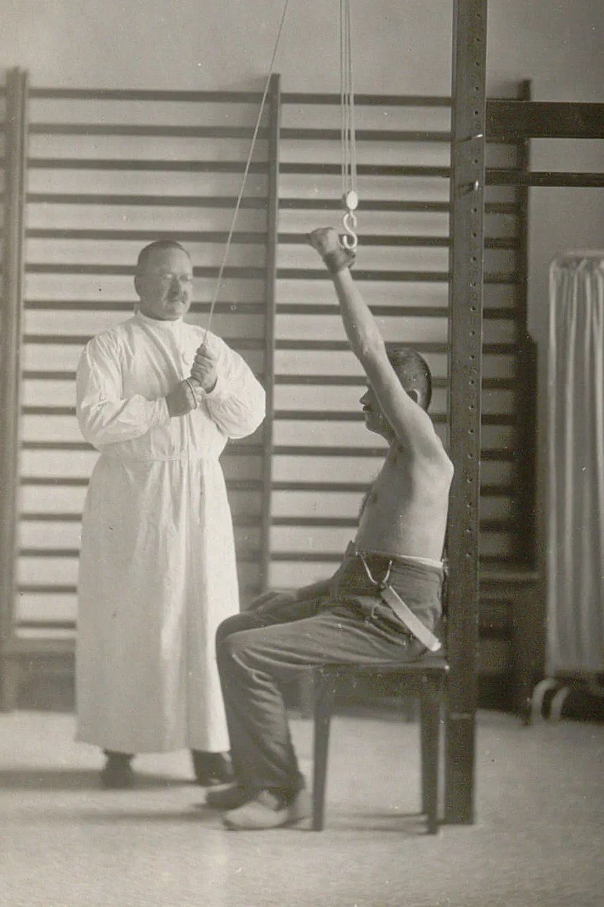 Homme à torse nu, assis sur une chaise, un physiothérapeute lui redresse les épaules grâce à un système de poulie.