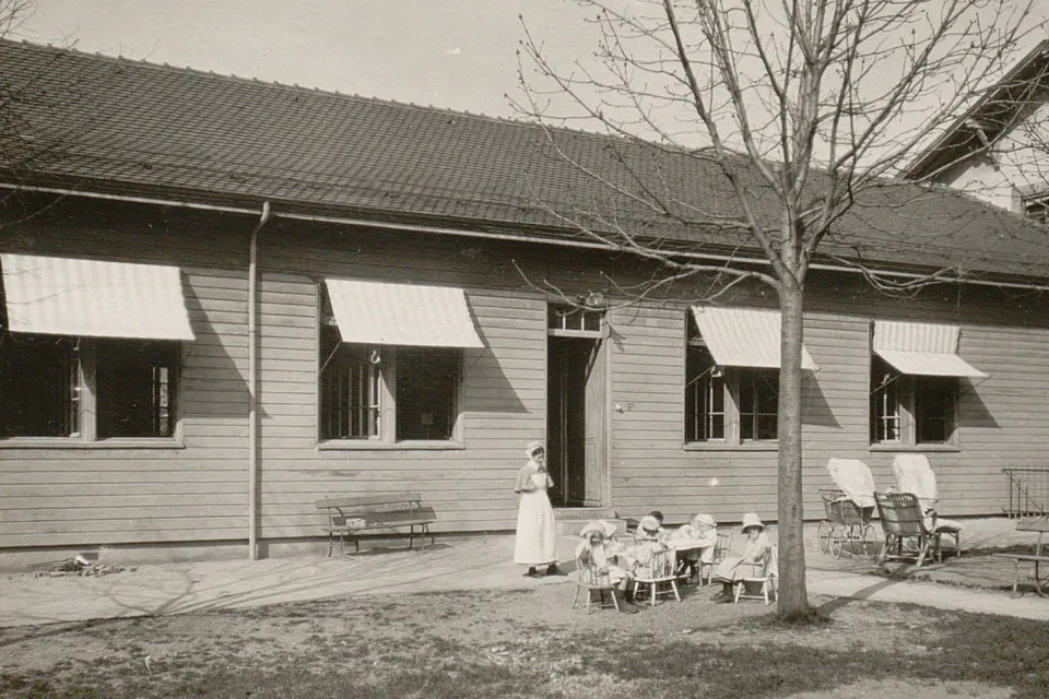Dans le jardin devant le pavillon, des jeunes enfants sont assis sur des petites chaises, surveillés par une infirmière.