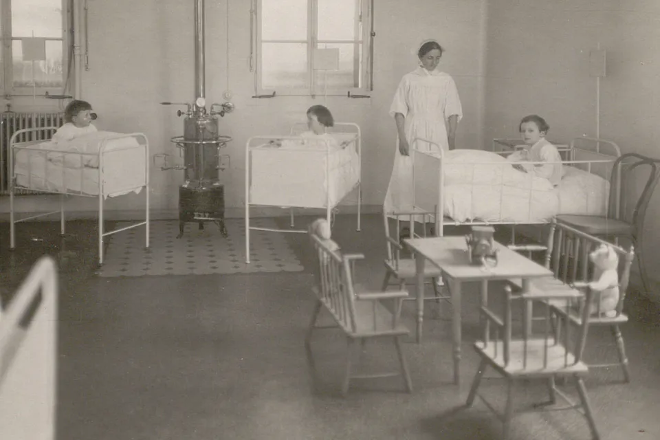 Trois jeunes enfants dans des lits à barreaux, surveillés par une infirmière. Un coin jeu avec un nounours.