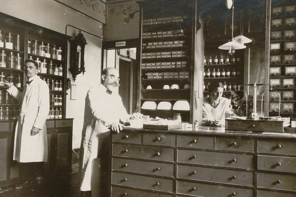 Trois personnes en blouse blanche devant des meubles remplis de pots, bouteilles et tiroirs.