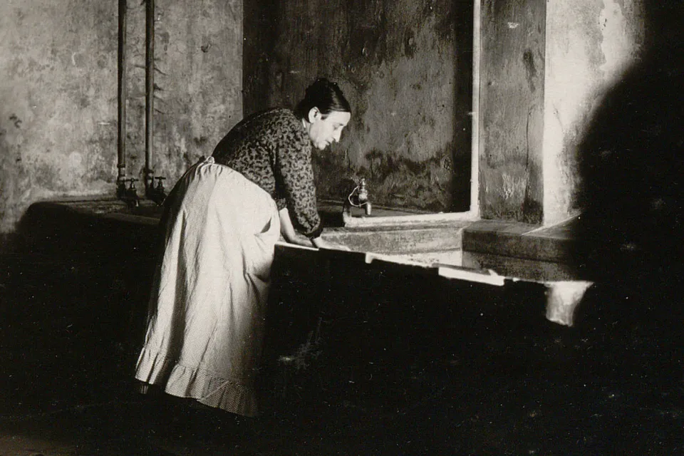 Une femme avec tablier se penche sur un lavoir muré.