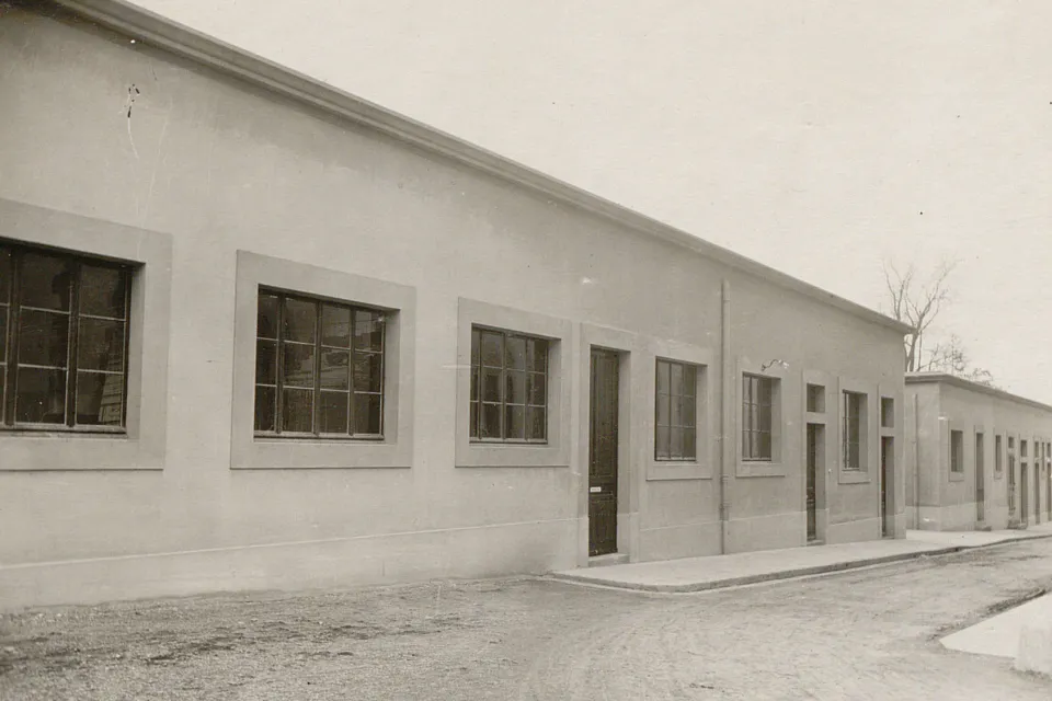 Une rangée de bâtiments bas au toit plat, avec des nombreuses portes et fenêtres sans volets, devant un chemin en terre.