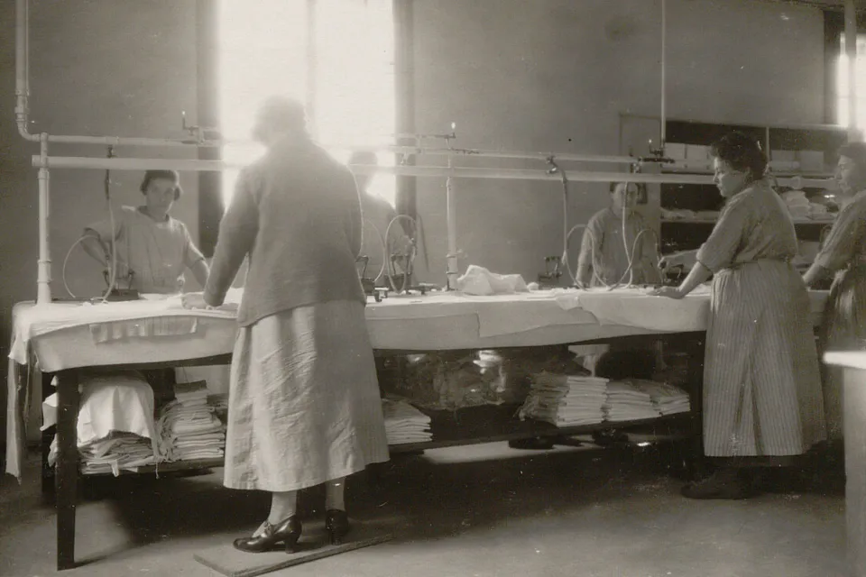 Six femmes autour d'une table couverte d'un molton, elles utilisent des fers à repasser à vapeur alimentés par des tuyaux.