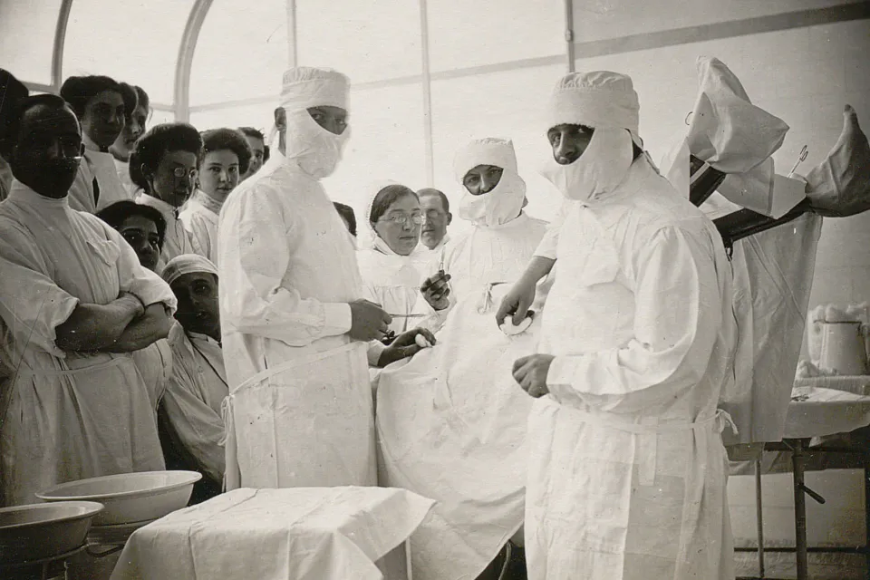 Trois chirurgiens avec charlotte et couvre-barbe ainsi qu'un public nombreux entourent un patient.