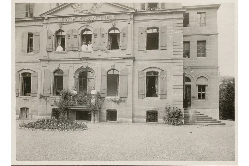 Bâtiment à plusieurs étages avec une inscription "Maternité". Des personnes en blouse blanche sont aux fenêtres.