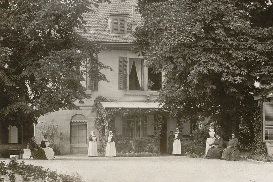 Une cour avec des grands arbres, plusieurs femmes sont assises sur des bancs. Des diaconesses sont près d'elles.