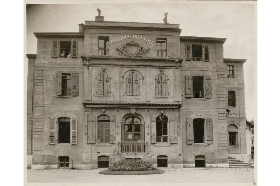 Bâtiment à plusieurs étages avec une inscription "Clinique infantile". Un balcon au centre, un escalier à droite.