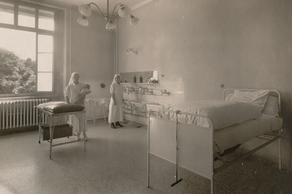 Une infirmière avec un bébé dans les bras dans une salle avec un lit d'hôpital très haut, des lavabos, un chandelier.