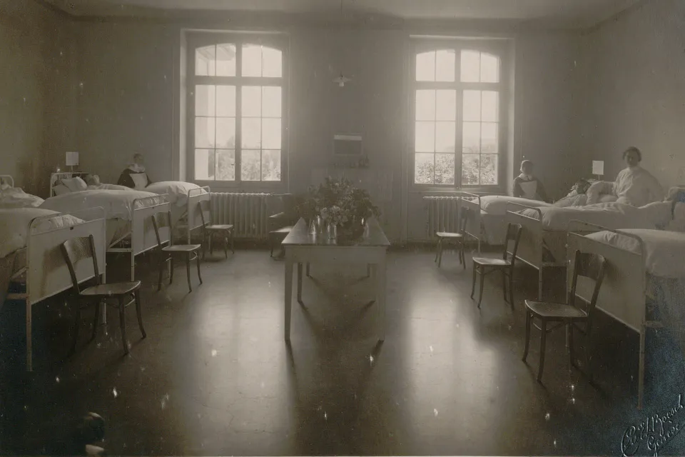 Chambre à six lits, plusieurs patientes et diaconesses, une table au centre avec un bouquet de fleurs.