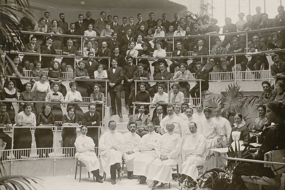 Un amphithéâtre rempli, en bas, le professeur et son équipe en blouses blanches.